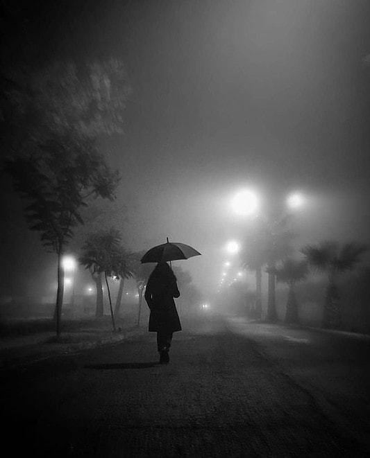 Black and white photograph of woman under a rain umbrella walking through fog under streetlights.