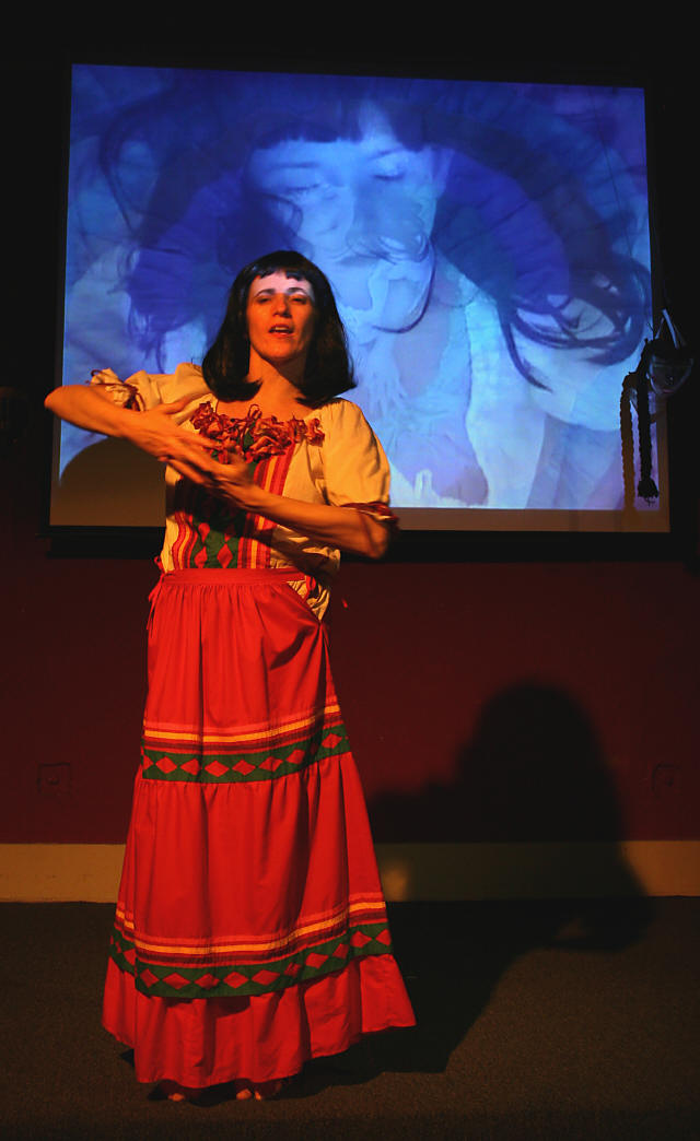 Aimee Greenberg in Dona Sangre holding her arms in a cradle position against an image of a young child in a projection.