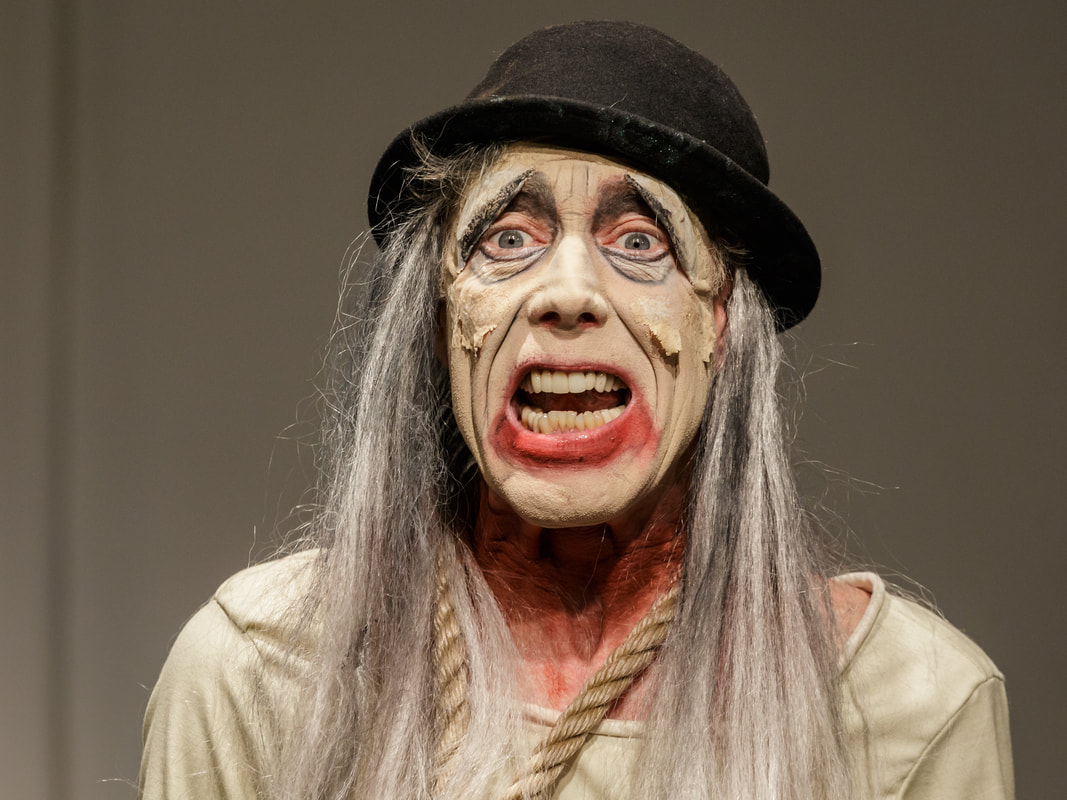 Lucky standing with a rope around his neck with long gray hair and red lips and peeling white make-up with an open-mouthed expression in Waiting for Godot.